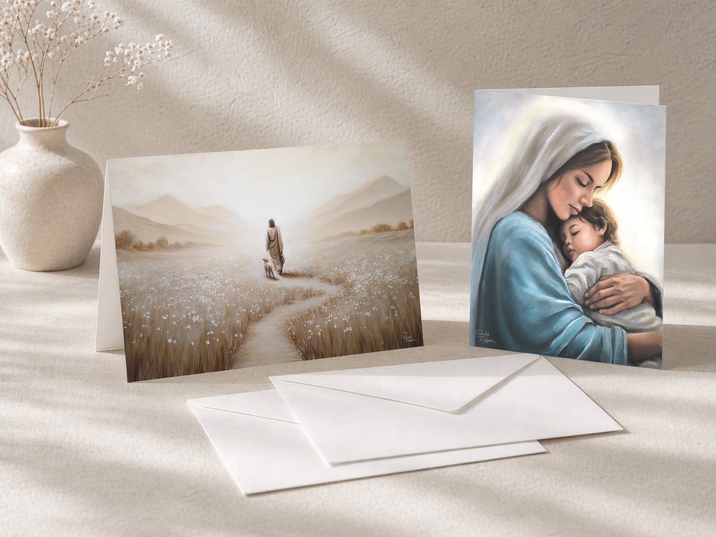 Two greeting cards on a table with envelopes, one featuring a couple in a field and the other showing Mary holding a child.