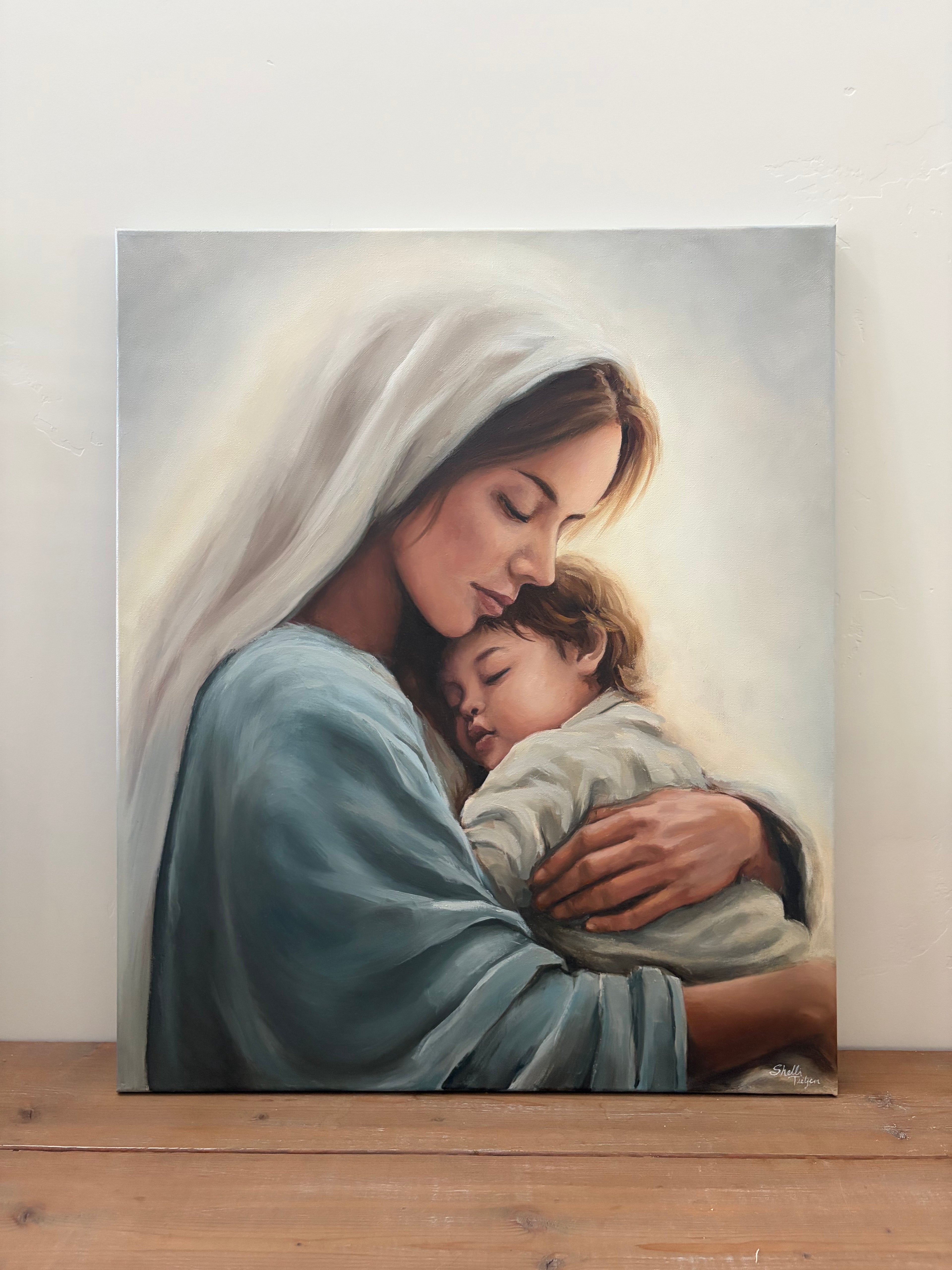 Picture of the original painting Held in Grace of Mary holding little Christ. Canvas is on a wooden surface with a white wall background