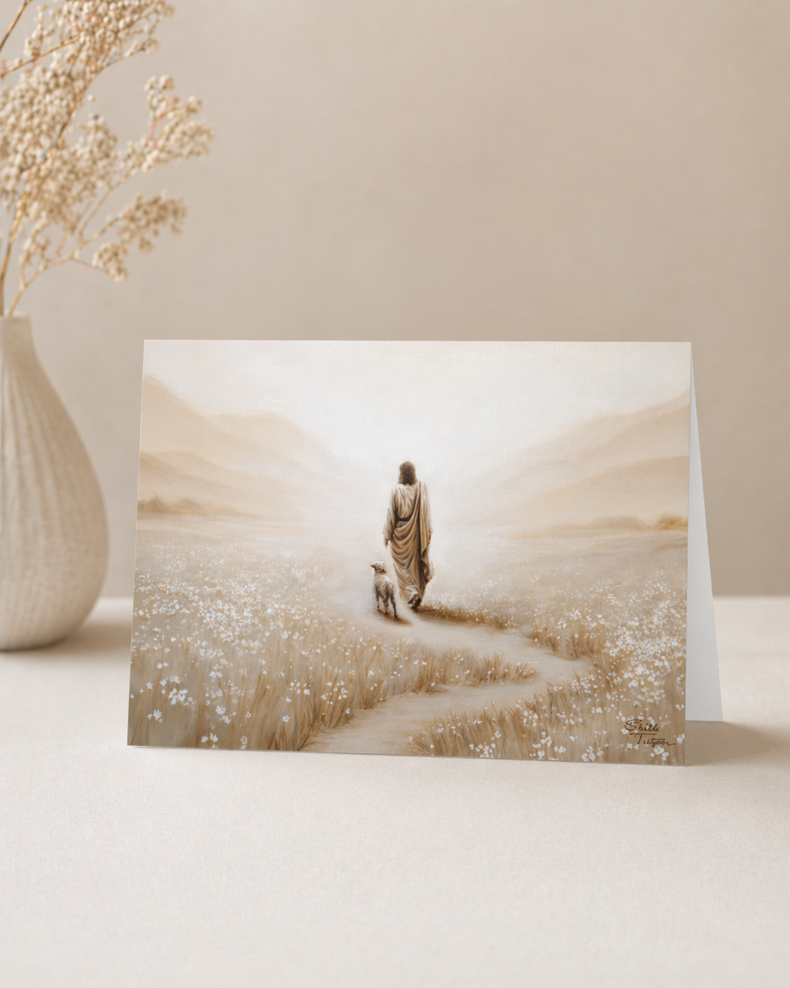 Greeting card with Jesus walking with a Lamb in a field, placed on a white surface with a vase in the background.